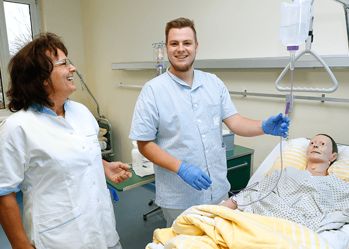 Ein junger Pfleger legt einer medizinischen Simulationspuppe eine Infusion. Neben ihm steht eine Ärztin, welche ihn lächelnd anleitet.