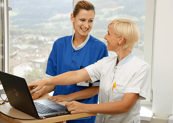 Eine Ärztin zeigt einer Pflegerin etwas auf dem vor ihnen stehenden Laptop. Beide blicken sich lächelnd an.